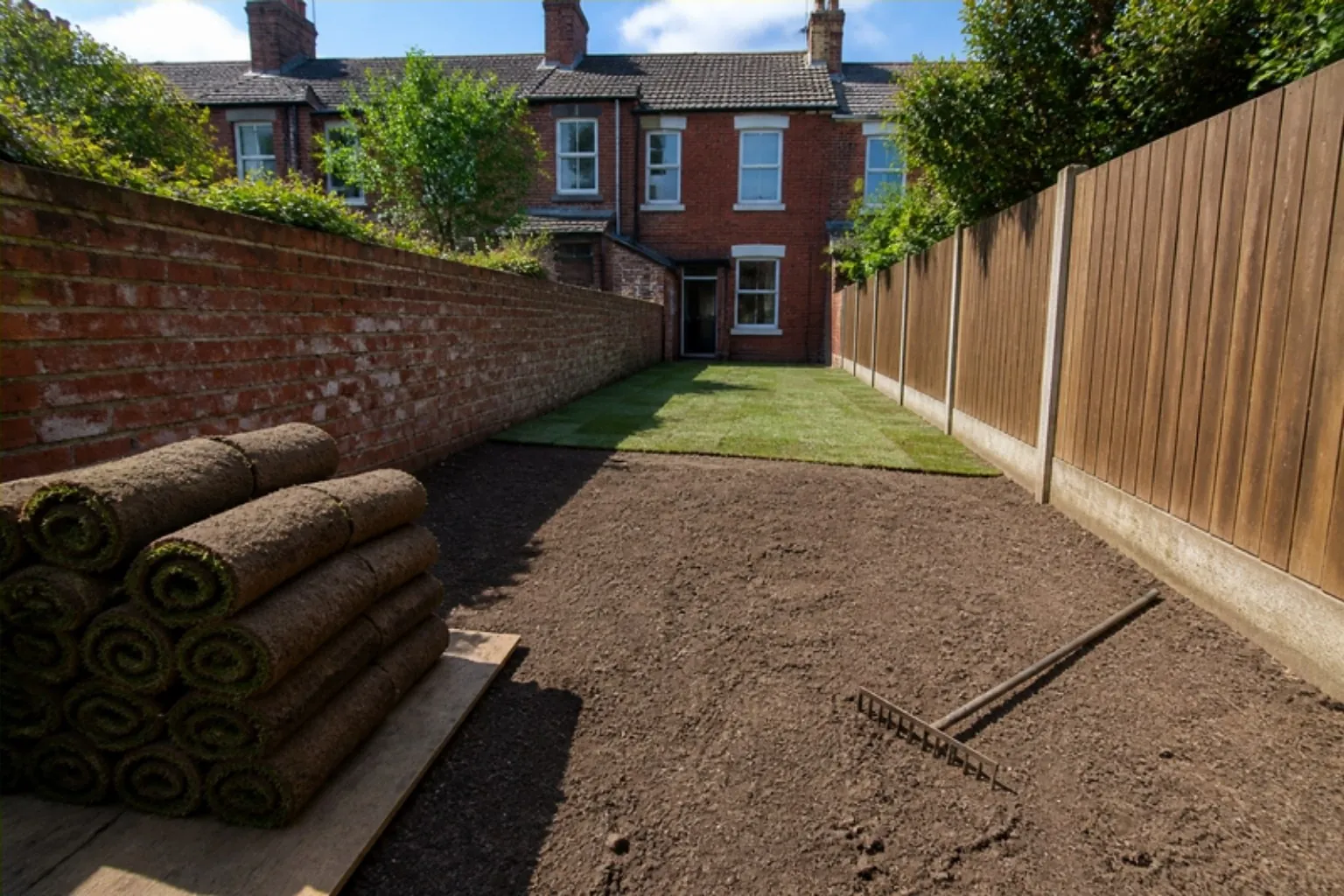 Narrow terrace garden half turfed, with levelled soil in front and rolls of turf stacked on a board