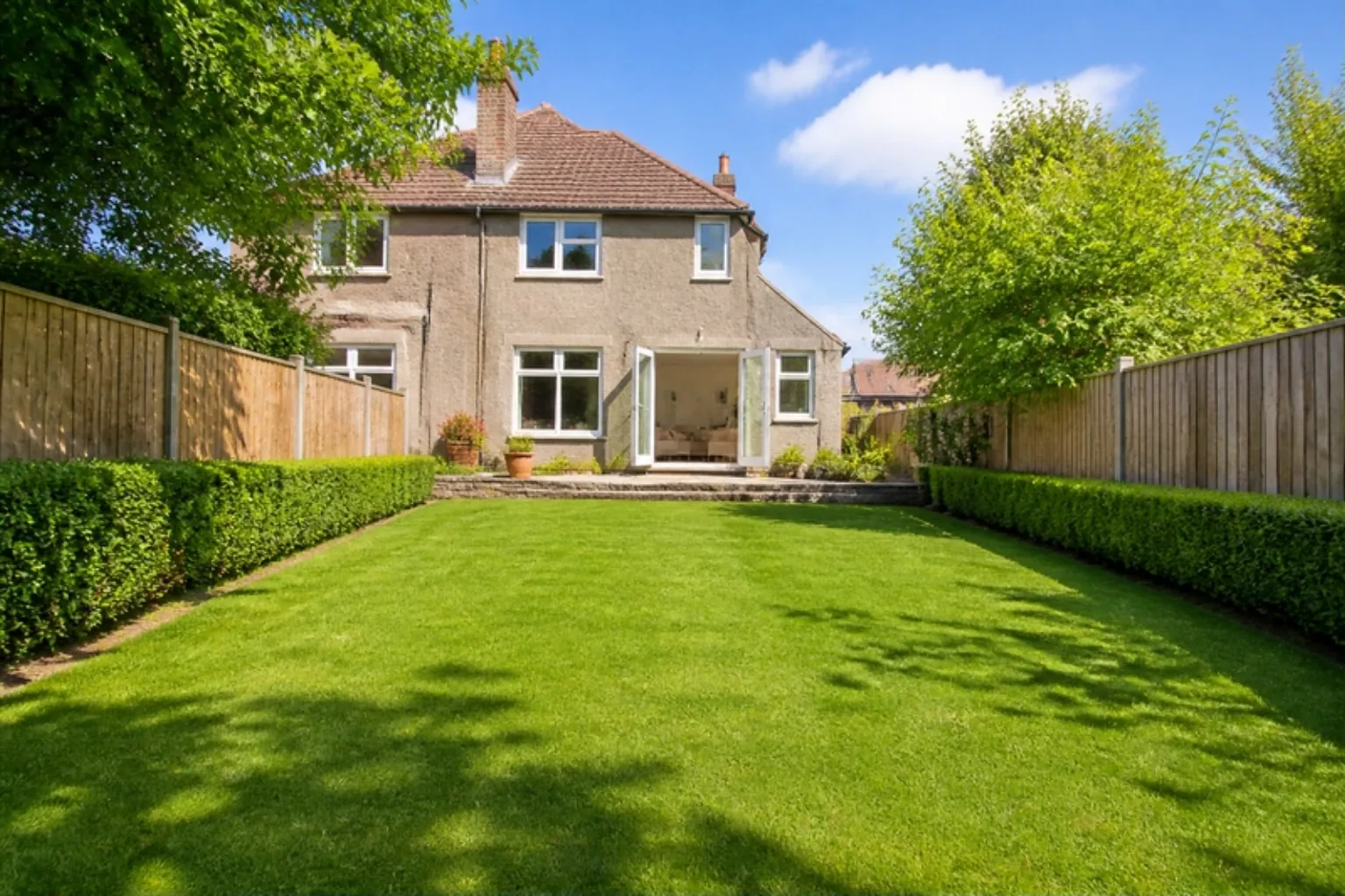 Long back garden of a 1930s pebbledash semi with a fresh green lawn, clipped hedges and open French doors
